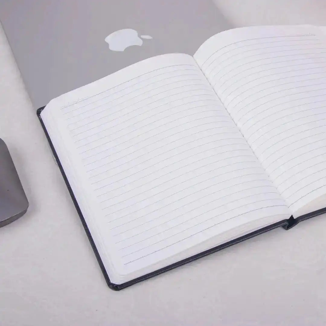 Black Primo A5 Diary with 80GSM lined pages open on a desk beside a laptop. Ideal for productivity planning and daily goal tracking.