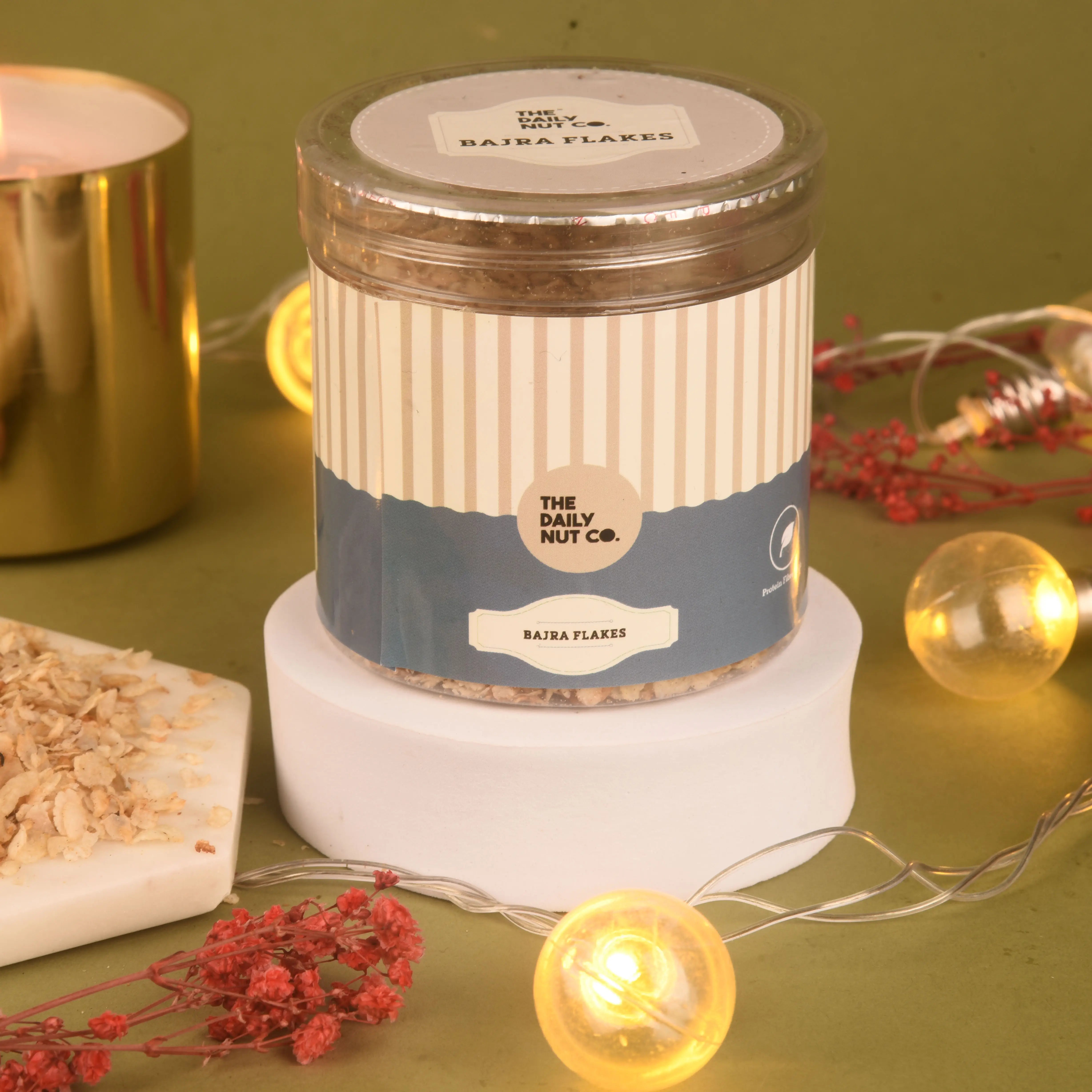 Jar of Bajra Flakes from The Daily Nut Co on a green surface with decorative elements.