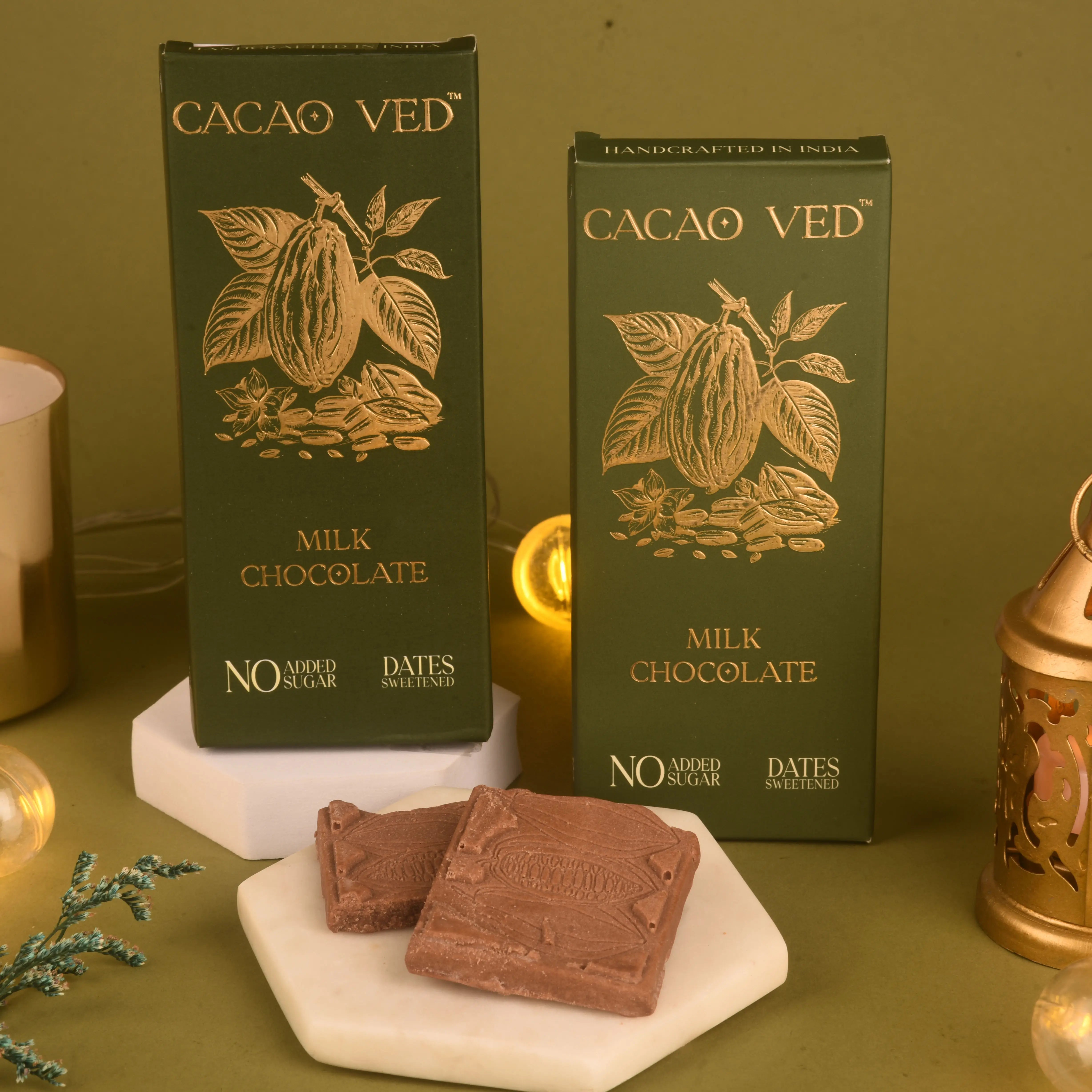 Two boxes of Cacao Ved milk chocolate on a green background with two pieces of chocolate in front.