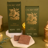 Two boxes of Cacao Ved milk chocolate on a green background with two pieces of chocolate in front.