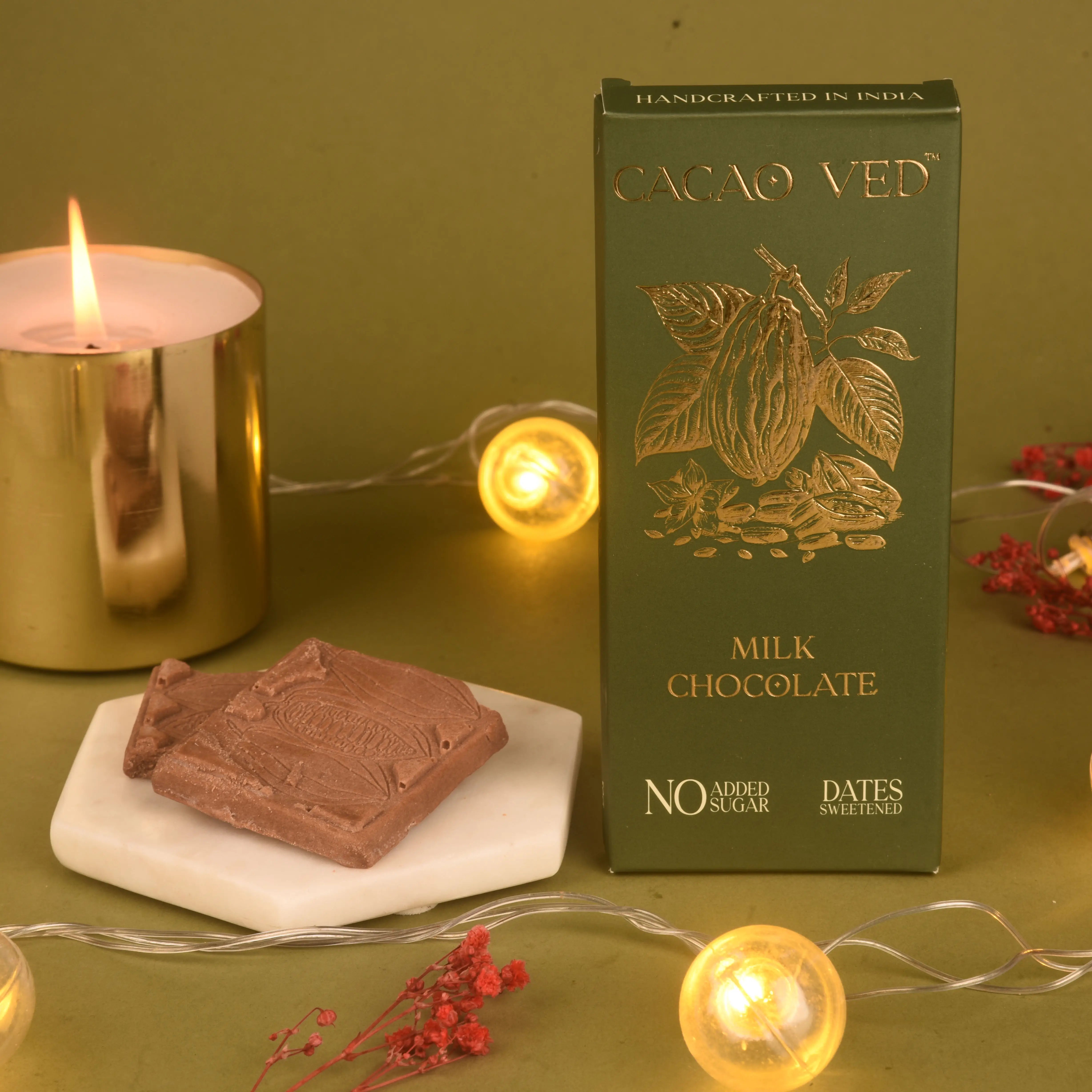 Cacao Vedi milk chocolate bar and packaging on a green surface with a candle and decorative lights.