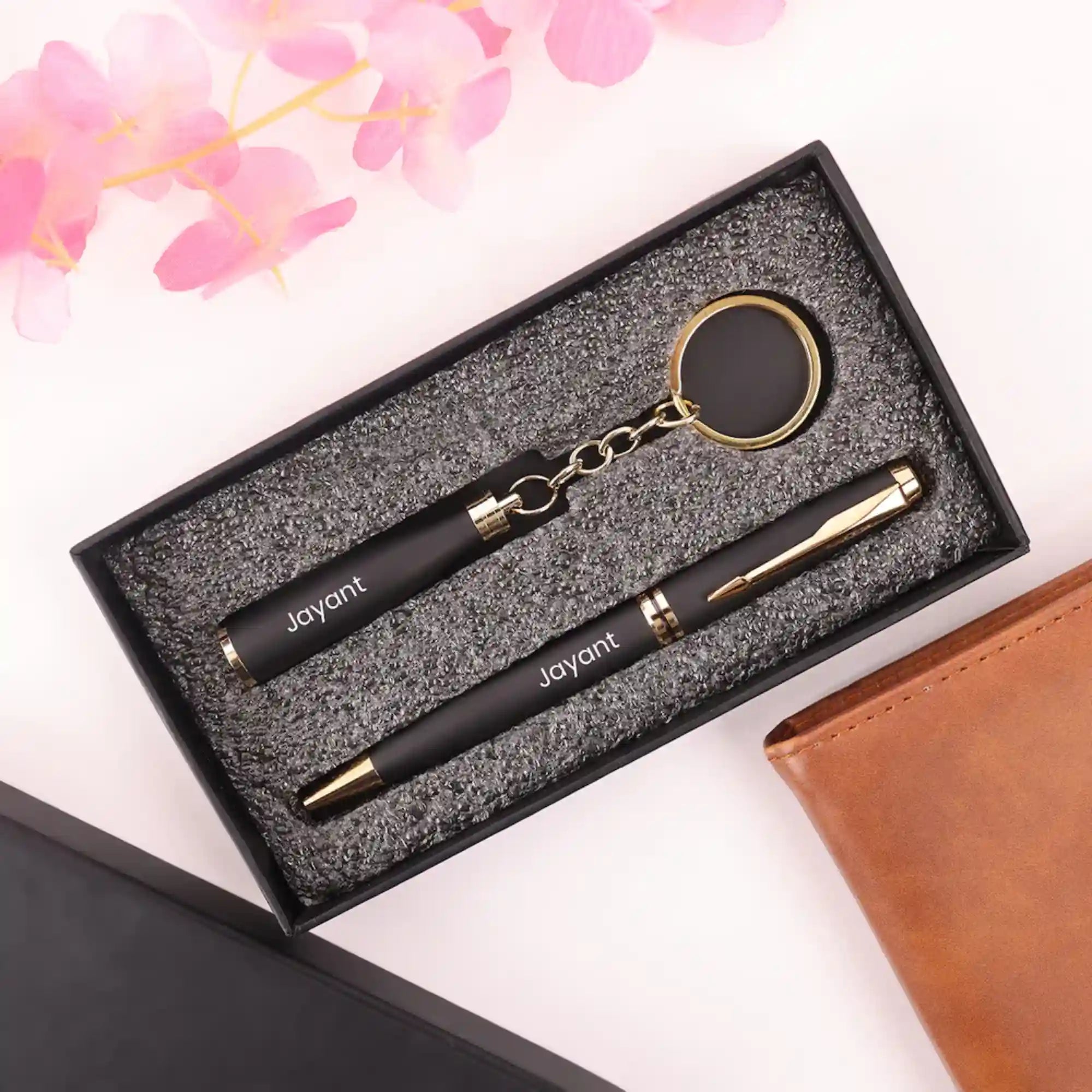 Black and gold pen set with keychain in a textured box, surrounded by pink flowers and a brown leather item.
