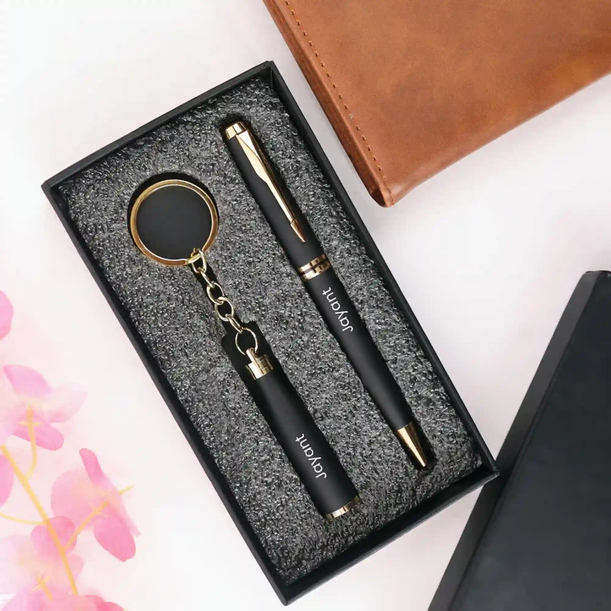 Black pen and keychain set with gold accents in a textured box, next to a brown leather notebook.