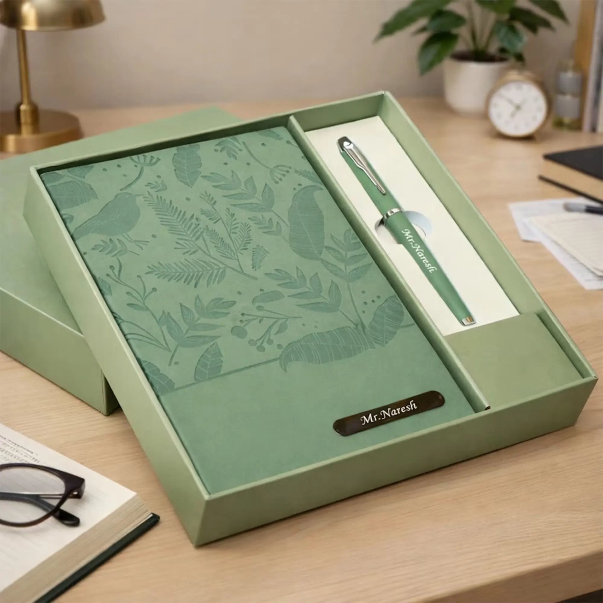 Green notebook and pen set in a box on a desk with a plant and clock in the background