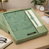 Green notebook and pen set in a box on a desk with a plant and clock in the background