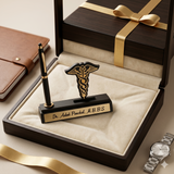 A high-angle photograph of a personalized luxury desk gift set resting on a beige velvet lining inside an open dark wood presentation box. The set includes a matte black base holding a black and gold pen and a black and gold metal medical caduceus (Hermes staff) symbol. A gold plaque on the front of the base is clearly engraved with "Dr. Ashok Panchal, M.B.B.S." Surrounding the box on a neutral surface are a leather journal, a silver wristwatch, and a gold ribbon.
