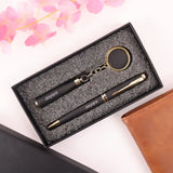 Black and gold pen set with keychain in a textured box, surrounded by pink flowers and a brown leather item.