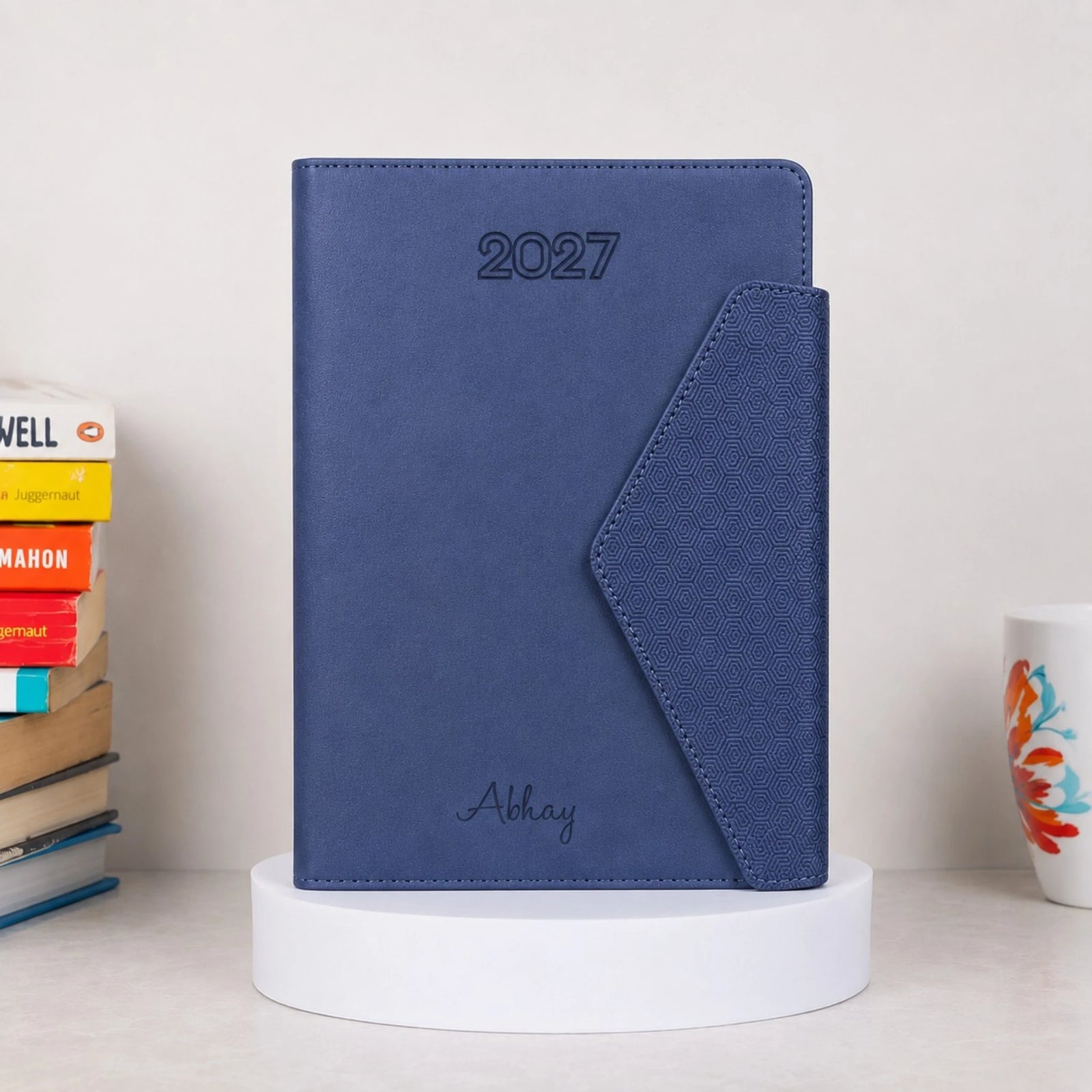 A blue textured 2027 planner or agenda with '2027' embossed at the top and 'Abhay' written at the bottom, displayed on a white pedestal. The planner is styled on a tabletop with books on the left and a mug on the right against a light background.
